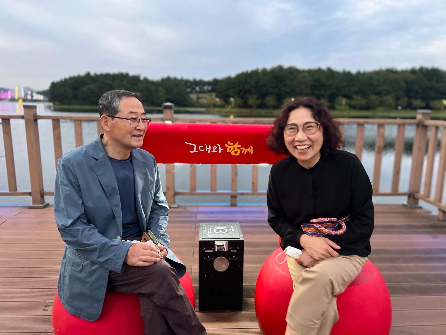 강변 데크 위 '그대와 함께' 의자에 나란히 앉아 웃는 두 사람